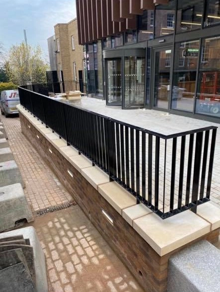  metal balustrade system, serving as a safety barrier outside a modern building entrance, treated with a durable, weather-resistant gloss black powder-coated finish to protect against the elements.Design Style: It features a clean, minimalist architectural style.